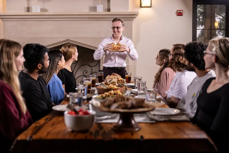Joseph Pool leading a conversation with students seated around a long table set for a shared meal